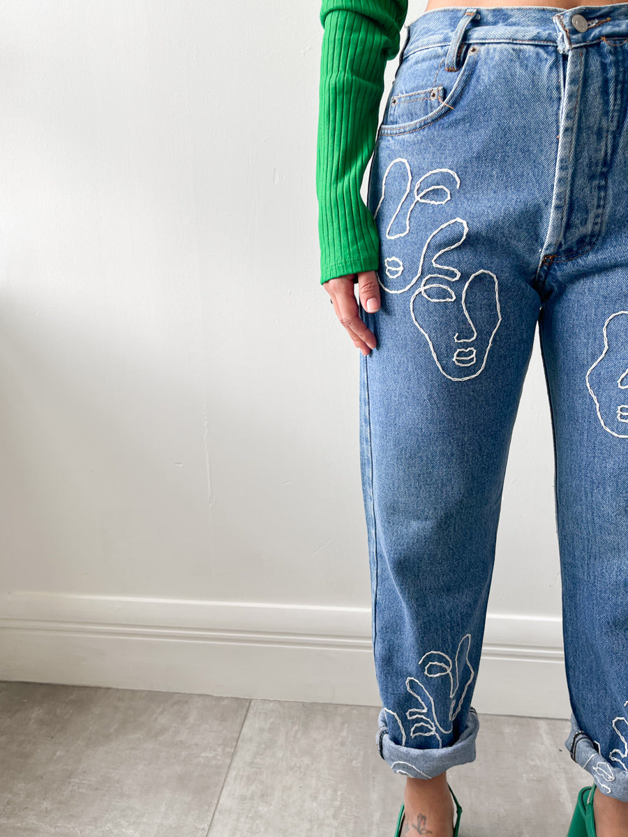 Fanfare Label sustainable women’s clothing brand UK. Our upcycled jeans are high-waisted, blue denim and made from recycled denim. We decorate every jean with a white embroidered thread in a face design.